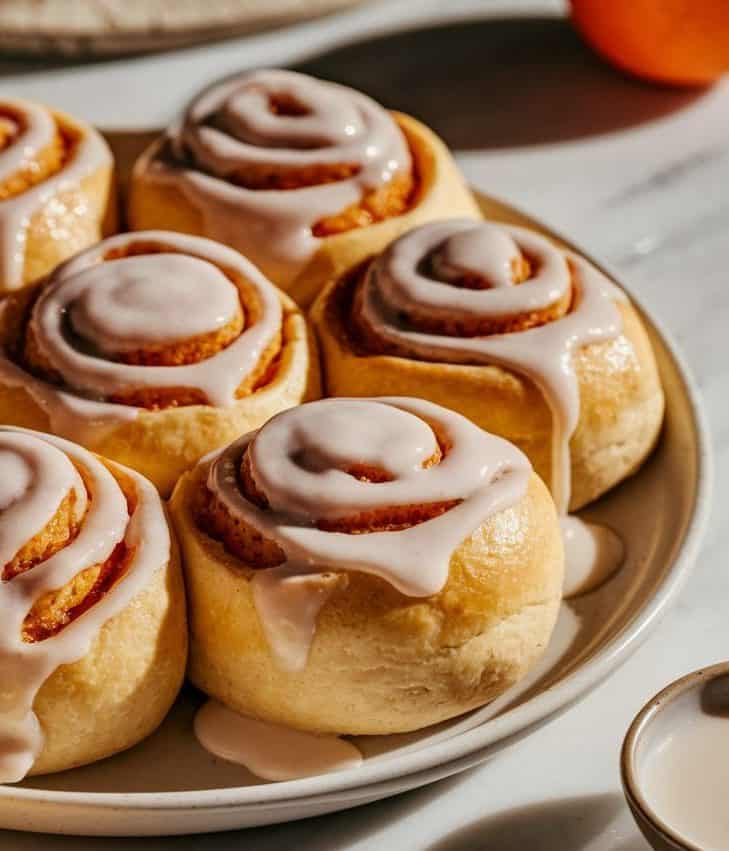 A close-up shot of soft, fluffy sourdough orange rolls on a large white ceramic plate, their plump bodies glistening with a rich, creamy white glaze that adds a lustrous sheen, revealing hints of cinnamon-orange zest filling. A small bowl of white glaze and a spoon rest beside the plate, while a blurred orange sits nearby, creating a warm, inviting atmosphere on a smooth white marble surface.