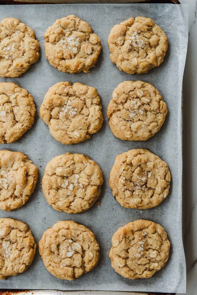A dozen of freshly baked sourdough oat cookies, elegantly arranged on a parchment-lined baking sheet on a white marble surface. The golden-brown cookies have a soft surface, with a little rock salt sprinkled in the middle.
