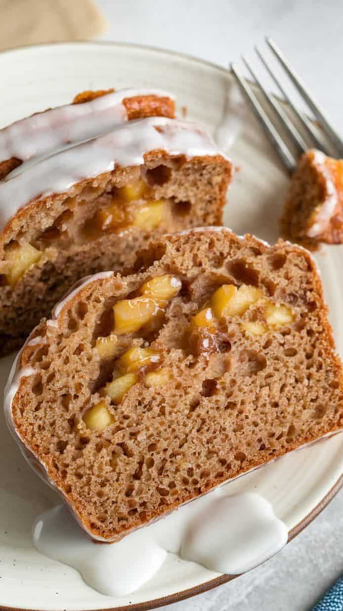 A close-up view of 2 slices of sourdough apple fritter bread loaf with white glaze on a white ceramic plate. The white glaze pools on the plate, adding a glossy, sweet finish. A few tiny minced apple pieces peek from the moist, soft, cake-textured brown crust at a few random tiny spots. The apple pieces are deeply caramelized, translucent, dark brown, and glossy. A fork rests beside the plate, with a bite-sized piece ready to be enjoyed.