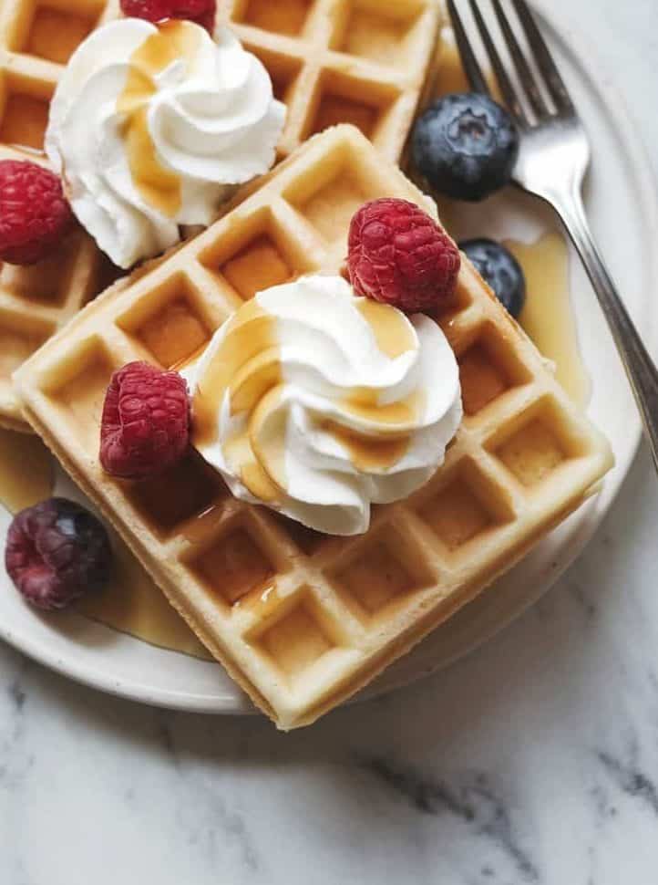 A top view of 2 freshly baked sourdough waffles on a white ceramic dish on a white marble surface. The waffles have a crisp, golden-brown exterior with deep squares. They are topped with a swirled dollop of whipped cream and a drizzle of maple syrup cascading down. Slices of fresh berries add color and texture. A fork rests beside the plate. The lighting is bright and natural, creating a welcoming atmosphere.