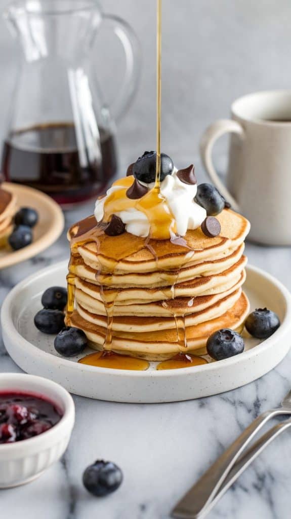 A stack of golden-brown sourdough discard pancakes beautifully arranged on a large white ceramic plate. Each pancake is generously topped with a dollop of fluffy whipped cream and a scattering of fresh blueberries and chocolate chips for added color and texture. A drizzle of warm maple syrup cascades down the sides, glistening under the light. On the side of the plate, a small white ceramic bowl filled with homemade berry compote complements the dish, inviting guests to indulge further. A fork with an elegant handle rests beside the plate, ready for enjoyment. In the background, a clear glass carafe filled with freshly brewed coffee stands next to a white ceramic mug, designed to enhance the overall cozy brunch atmosphere. The setting is on a clean, white marble surface, providing a stunning contrast to the vibrant colors of the pancakes and toppings, ensuring a visually appealing presentation that tempts the eyes and the palate alike.
