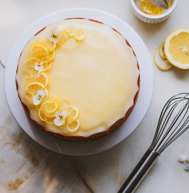 A freshly baked sourdough lemon cake with lemon glaze and lemon toppings, served on a ceramic plate and surrounded by lemon slices, a whisk, and a small ceramic bowl of lemon juice